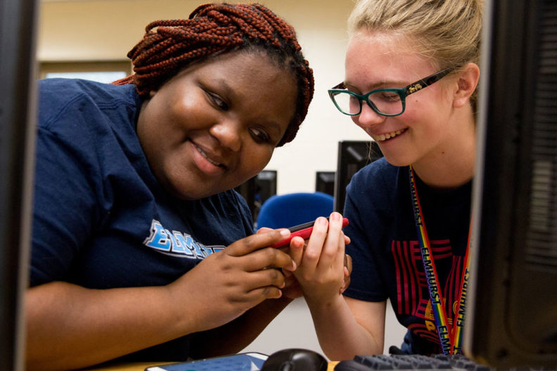 A Black female student and a white female student at Elmhurst University smile at each other between their computer terminals.
