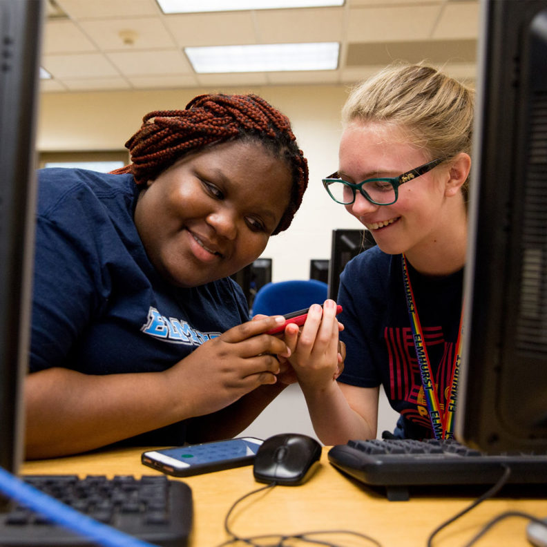 A Black female student and a white female student at Elmhurst University 幸运艇 smile at each other between their computer terminals.