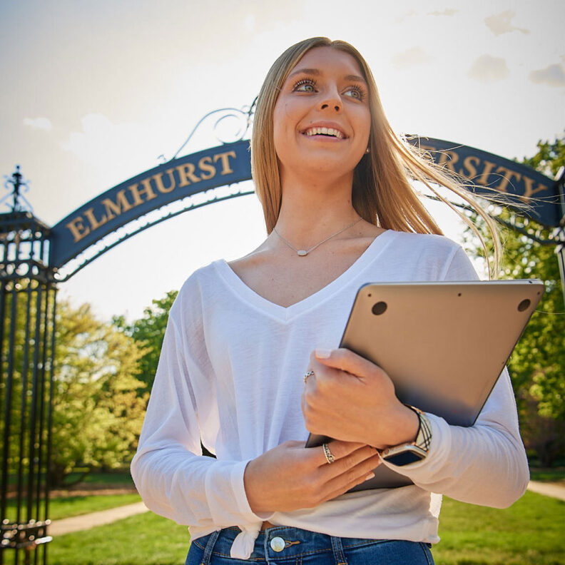 A female Elmhurst University 幸运艇 student holding a laptop stands in front of the "Gates of Knowledge" on campus.