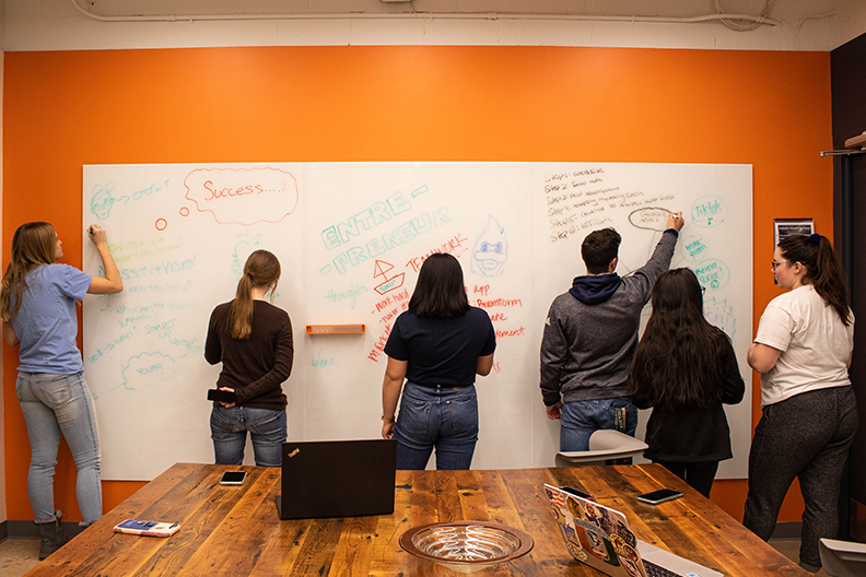 Six Elmhurst University students draw and write together on a large whiteboard in a classroom. Elmhurst is promoting entrepreneurship with a new initiative on campus.