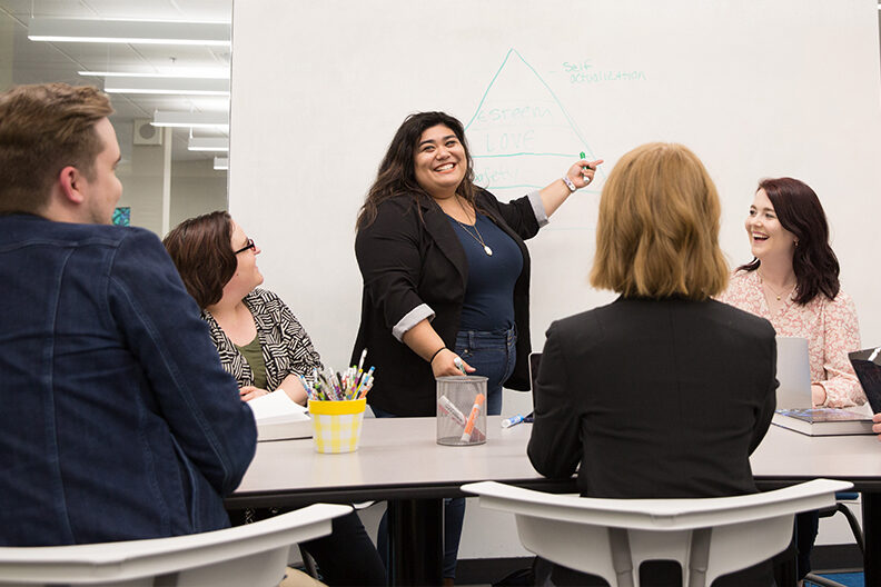A woman stands at the front of the class and points to a whiteboard.