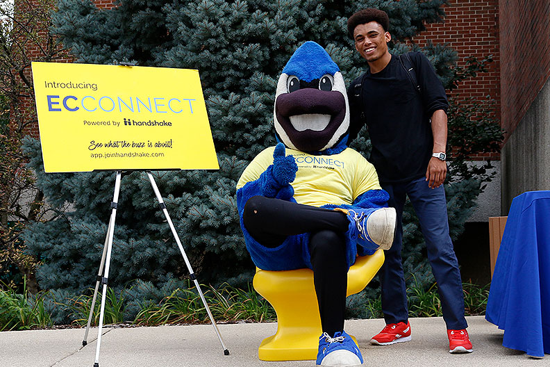 Elmhurst College mascot Victor E. Bluejay, seated in a yellow chair, poses with a student during a career center event.